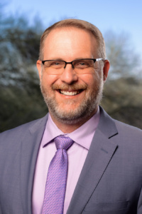 Profile pic of John Sparks - white bearded male wearing glasses and a gray suit with purple shirt and tie in front of mesquite tree background