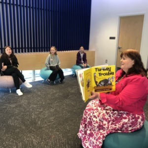 Teacher holding a book reading to three students sitting in chairs