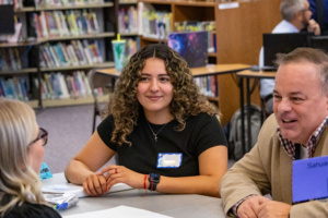 Female teacher with curly hair wear b lack shirt smiling. Male teacher to the right wear brown blazer